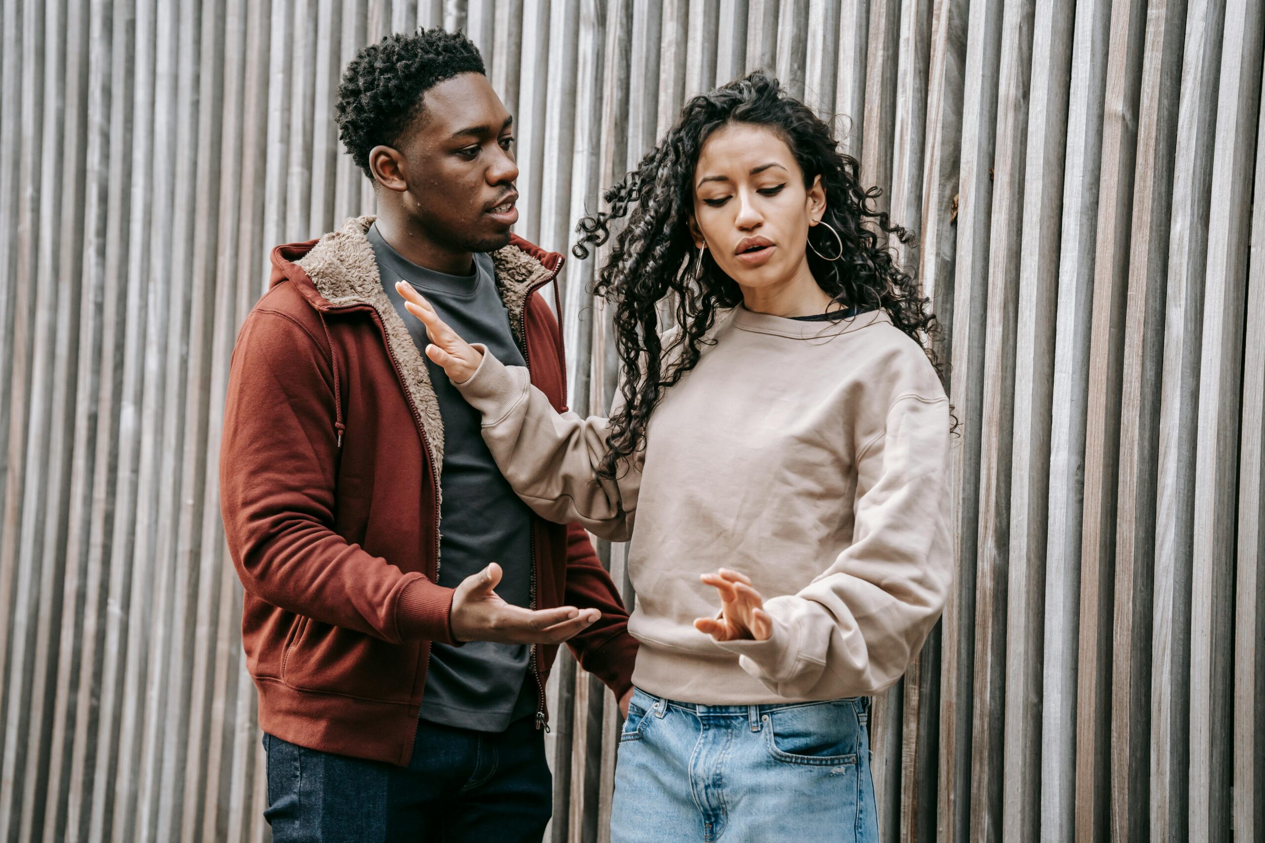 A diverse couple expressing conflict and disagreement outside against a wooden fence.
