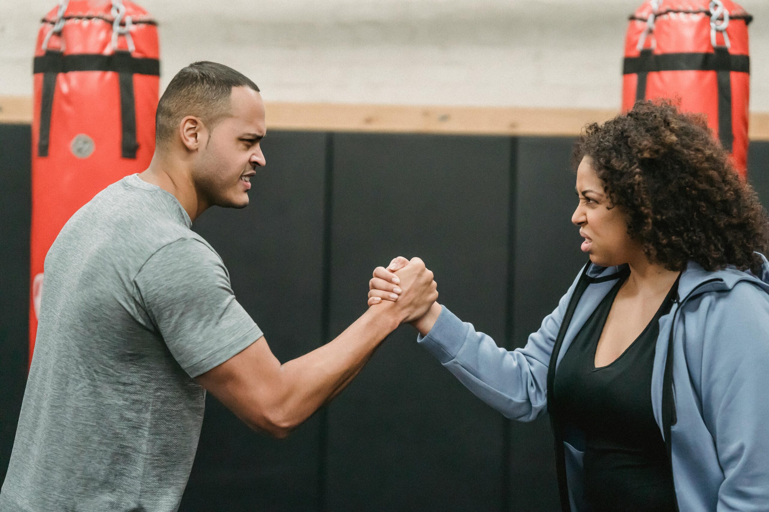 Side view of angry African American couple clasping hands and looking at each other while showing strength during workout in boxing gym