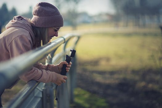 Woman in a beanie using a camera on a stabilizer, leaning on a railing outdoors during the day.